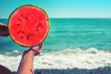 Round watermelon, juicy slice in the form of the sun, in the hands of a woman against the sea, in the rays of sunlight. Diet of tropical fruits..