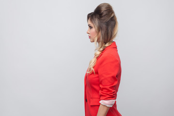 Profile side view portrait of serious beautiful business lady with hairstyle and makeup in red fancy blazer, standing and looking straight. indoor studio shot, isolated on grey background.