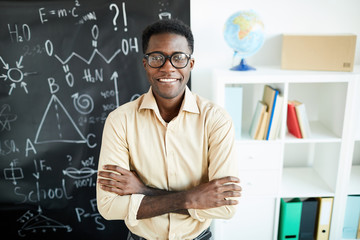 Young successful African-american teacher with arms crossed on chest standing by blackboard in...
