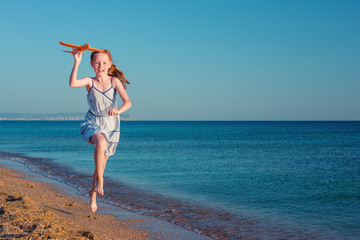 Fototapeta na wymiar Girl runs along the beach. Happy red-haired child, playing with a toy airplane, runs along the sandy beach of the sea against the blue summer sky.