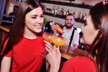 two young cute girls drink cocktails in a nightclub or bar, have fun, smile and talk to the bartender
