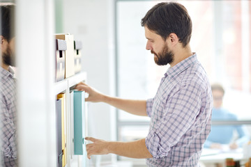 Bearded businessman taking one of folders with archive data from shelf while looking for some...
