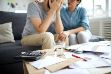 Fototapeta premium Glass of water, some payment bills or finacial papers, calculator and other stuff on table on background of young woman supporting her stressed husband