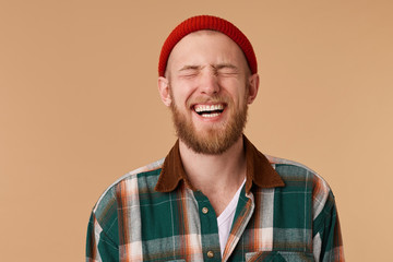 Laughing man in checkered shirt and red hat over beige wall. Toothy smile and beard. Male laughing out loud at funny meme he found on internet. Positive human facial expressions and emotions