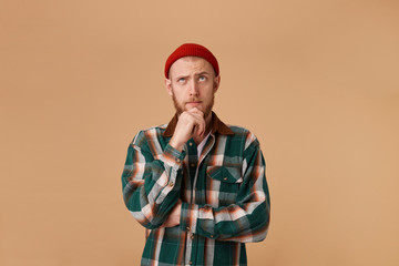 Forgetful confused curious bearded man in checkered shirt and hat, holding fist on chin and with thoughtful clueless expression, looking right upper corner, isolated over beige background.
