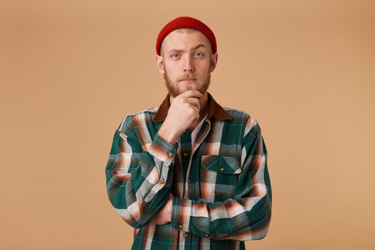 A confident man with a beard dressed in cool red cap and a checkered shirt holds his hand on chin, one eyebrow playfully raised thinks about something, build a plan, ponders a cool idea.