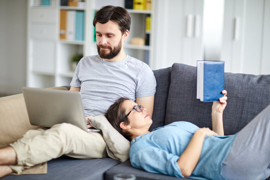 Relaxed spouses reading and networking on sofa while staying at home on weekend
