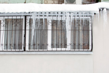 Dangerously hanging icicles from the roof over the window with a fence in winter. Sharp icicles on the roof of the house on the street hanging in a row