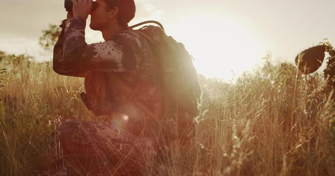 Orbit shot of crouched bowhunter looking through his binoculars
