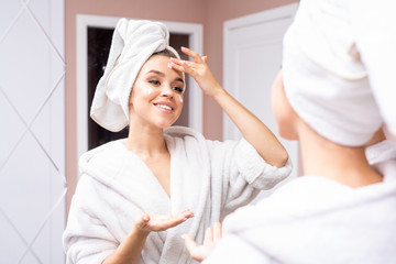 Portrait of beautiful young woman wearing bathrobe applying face cream while looking in mirror...