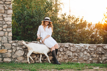.A girl in a white dress walks with a goat in nature.