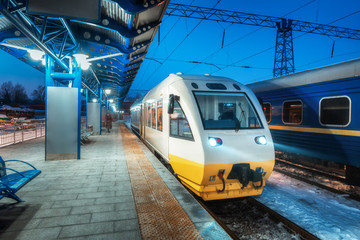 Fototapeta premium High speed train on the railway station at night in Europe. Urban landscape with modern commuter train on the railway platform with illumination at dusk. Intercity vehicle. Passenger railroad travel