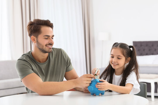 Little girl with her father putting money into piggy bank at table indoors