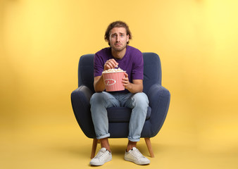 Emotional man with popcorn sitting in armchair during cinema show on color background