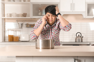 Emotional man calling plumber near table with saucepan under leaking water from ceiling in kitchen