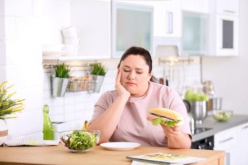 Sad overweight woman holding sandwich at table in kitchen. Healthy diet