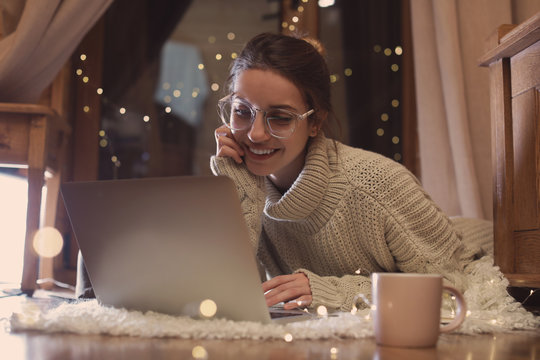 Woman with cup of hot beverage using laptop at home in winter evening