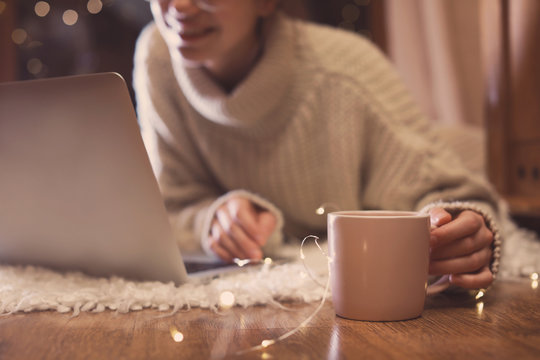 Woman With Cup Of Hot Beverage Using Laptop At Home In Winter Evening, Closeup