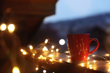 Cup of hot beverage on balcony railing decorated with Christmas lights, space for text. Winter...