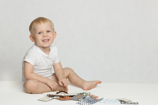 A toddler boy sits, laughs and plays with pills on a white background.