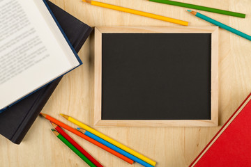 Black, empty, blank chalkboard with colored pencils and books on brown wooden desk flat lay from above