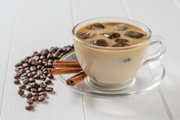 Glass bowl with ice coffee, cinnamon sticks and scattered coffee beans on white table.