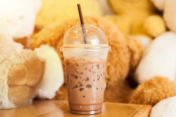 Cocoa cold with ice in the cup with straw put it on the wood table with bear doll in the cafe. - Image 