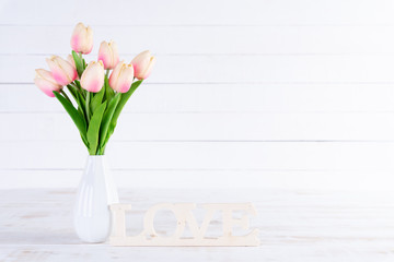 Valentines day and love concept. Pink tulips in vase with Wooden letters forming word LOVE written on white wooden background.