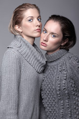 Two young women in gray sweaters on grey background. Beautiful girls stretching hands forward in embrace. Female friendship concept