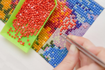 Girl collect diamond painting. Diamond embroidery with a tweezers. Acrylic rhinestones. Closeup,...