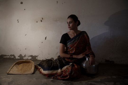 Beautiful Village Woman Sitting On The Floor With A Steel Pot And Winnowing Fan In An Old Low Lit Room Looking Away.	