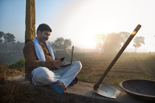 Happy looking farmer sitting near his agriculture field with a spade and iron gold pan by his side making online transaction on laptop computer using credit card.	
