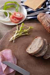 Freshly baked bread cut in slices on wooden chopping board with vegetable, cheese and ham on wooden table as background