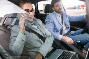 Young business people working together while traveling by a car. Businesswoman talking at phone while businessman driving car.