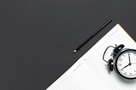 Flat lay black alarm clock, clean open notebook diary, pencil on gray dark background top view copy space. Concept time to work, desktop, Office desk, Minimalistic background for blog