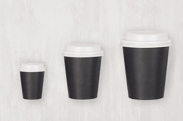 Coffee mockup - set of three different size black paper cups and blank white caps on white wood board, top view. Modern elegant concept for branding identity, advertising, design.