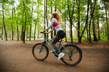 Fototapeta na wymiar Active girl in sportswear sitting on bicycle while moving down forest road on summer day or weekend