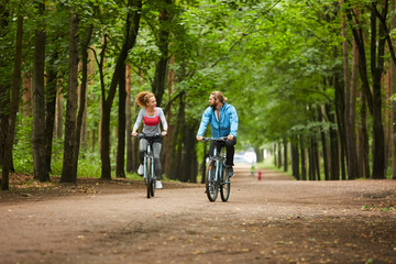 Young active man and woman sitting on bicycles while moving along road in park and talking to one another
