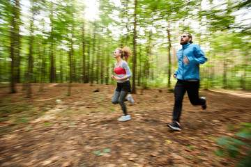 Active young couple in sportswear running down forest road on warm summer morning with blurry trees on background
