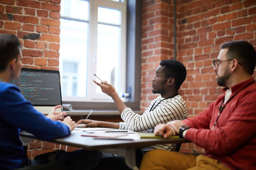Three young intercultural it-engineers sitting by table in front of computer monitor and discussing...