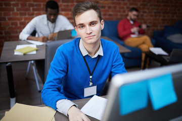 Young contemporary office manager sitting by table in front of computer monitor and camera during work