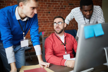 Two young intercultural office managers listening to their boss while looking at computer screen by...