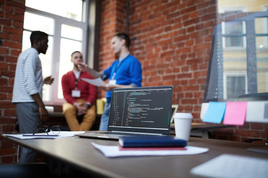 Technical algorithm or chain of codes on laptop display among other supplies on table and group of computer engineers on background