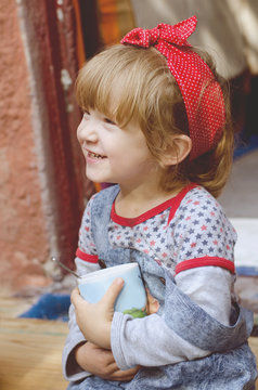 White cute toddler girl wearing bright red bandana on a had and retro style denim drinks cocoa while sitting on a house threshold. Happy child portrait.