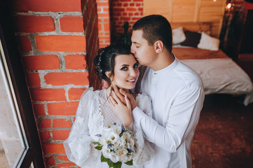 Stylish groom in a white shirt and a beautiful bride in a lace dress are standing near a brick wall in the studio. Wedding portrait. Wedding photography.