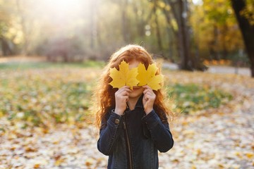 Autumn vibes, child portrait. Charming and red hair little girl looks happy walking and playing on the fallen leaves in autumn park full of evening sunshine