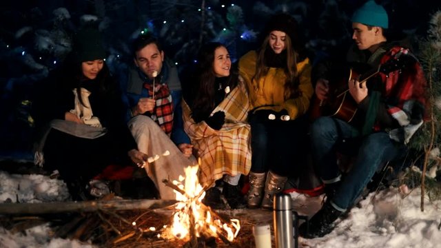 Group of friends sitting in winter forest by the fire, eating marshmallows and singing songs