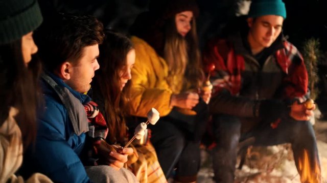 Group of young people in the winter forest sitting by the fire. Warming up marshmello by the fire