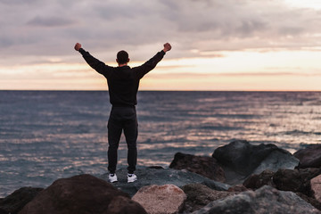 Fototapeta na wymiar Rear view of a happy unidentified man athlete rejoices in a fulfilled dream by raising his hands up and looking at sunset by the sea on warm summer evening. Concept of achieving goals and results