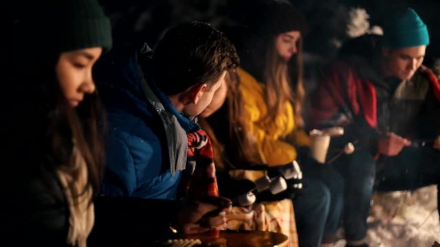 Young people in the winter forest sitting by the fire. Warming up marshmello by the fire